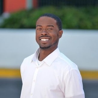 A smiling man in a white shirt standing outdoors with a blurred background that suggests an urban setting.
