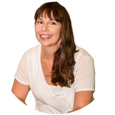 A smiling woman with long hair and bangs, wearing a white v-neck top, posing against a plain background.
