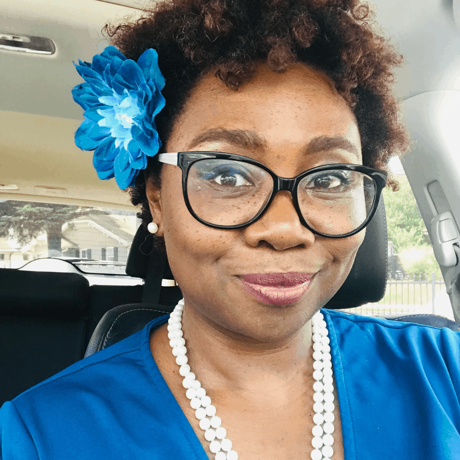 A woman with a bright smile, wearing glasses, and a blue flower in her hair, dressed in a blue top with a white necklace, taking a selfie in a car.