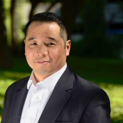 A confident professional man dressed in a suit without a tie, standing outdoors with trees in the soft-focus background.