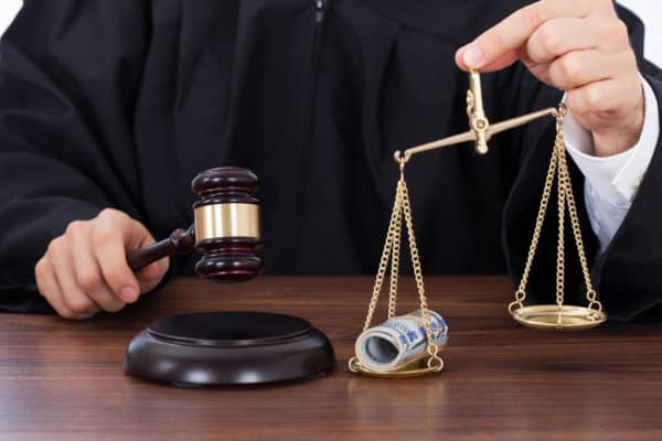 Midsection of male judge striking gavel while holding scale with money in courtroom