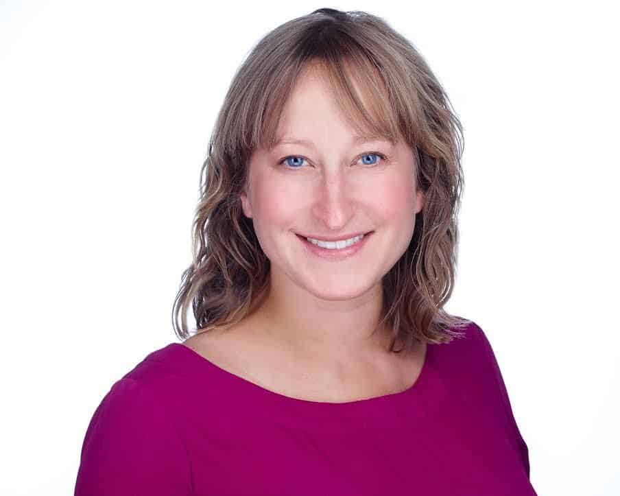 A smiling woman with shoulder-length hair wearing a purple top against a white background.