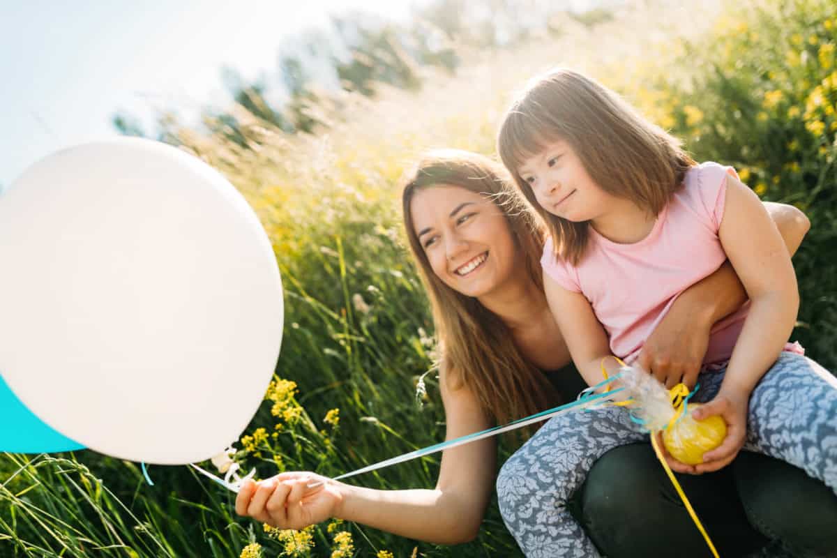 Mother and daughter enjoying a sunny day outdoors with a white balloon, sharing a moment of happiness in a field of tall grass.