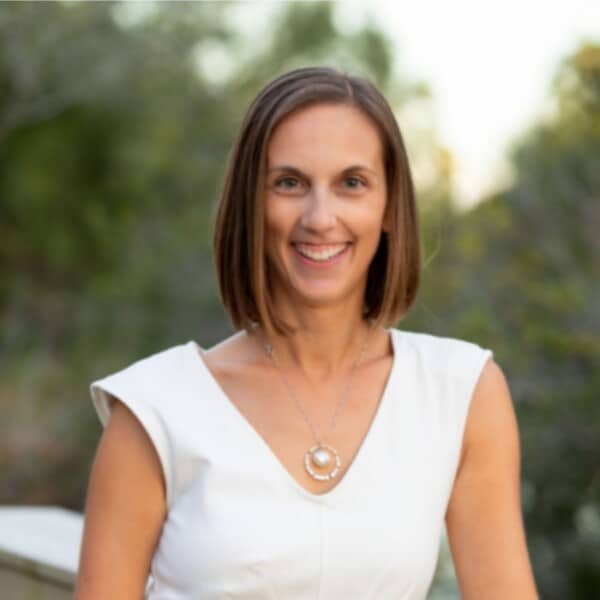 A smiling woman with shoulder-length hair, wearing a white top and a circular necklace, with a blurred green background suggesting an outdoor setting.