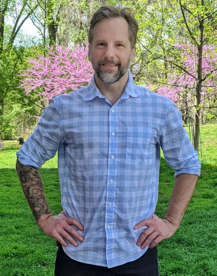 A man with a warm smile, sporting a blue plaid shirt and a visible tattoo on his left arm, stands confidently against a serene backdrop of green grass and blooming pink trees on a bright sunny day.