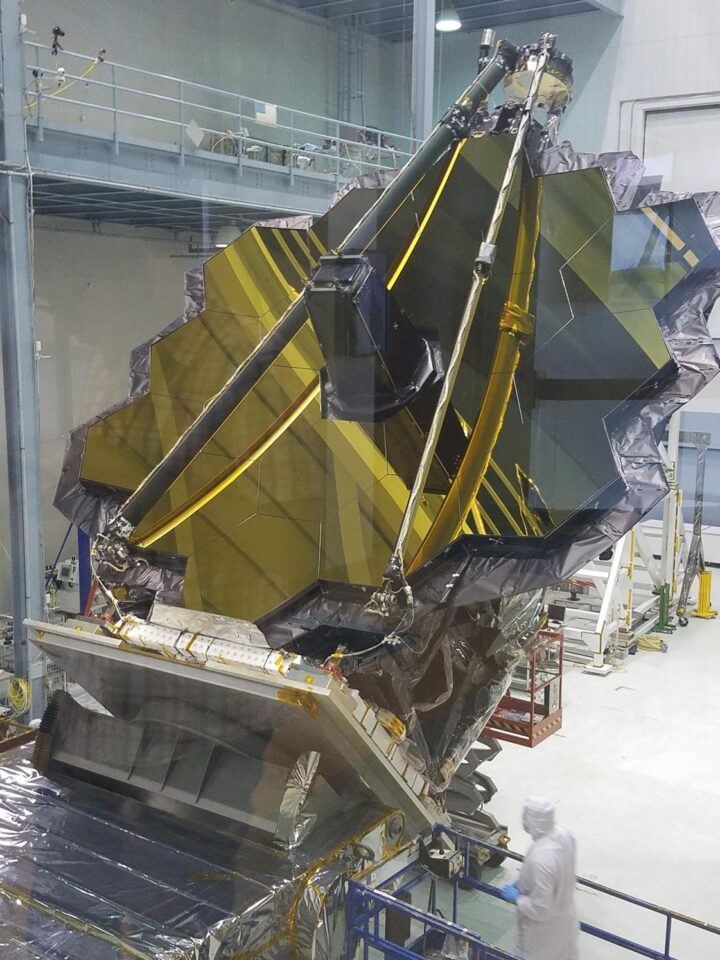 A team of technicians in a clean room working on the intricate assembly of a large, hexagonal, gold-coated mirror segment of a space telescope.