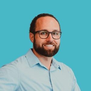 A professional man with a beard, wearing glasses and a blue shirt, smiling against a blue background.