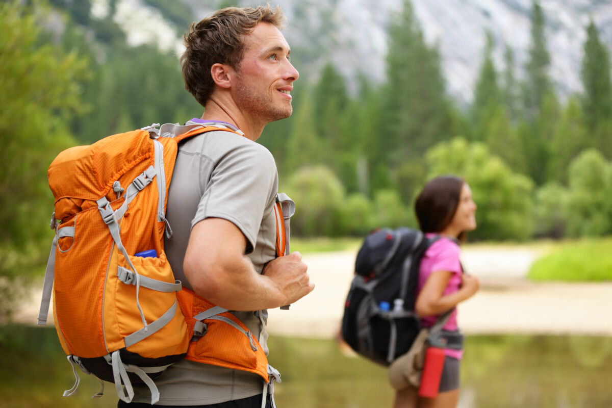 A male hiker with an orange backpack looks out over a tranquil natural landscape, while a female companion stands in the background near a body of water, surrounded by lush greenery and mountains.