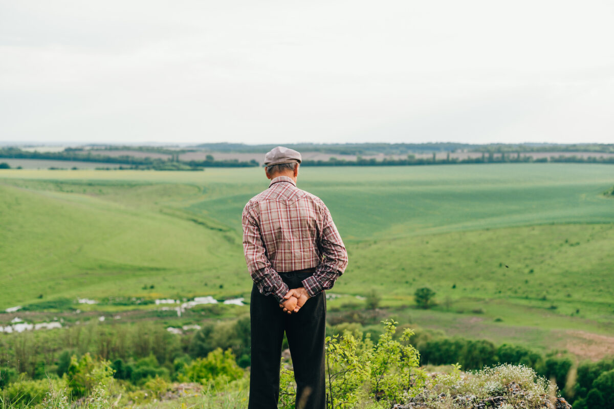 A retired man in a shirt and cap stands on top of a mountain with a beautiful green landscape. Photo of the grandfather of the pensioner from a back on a mountain walk.
