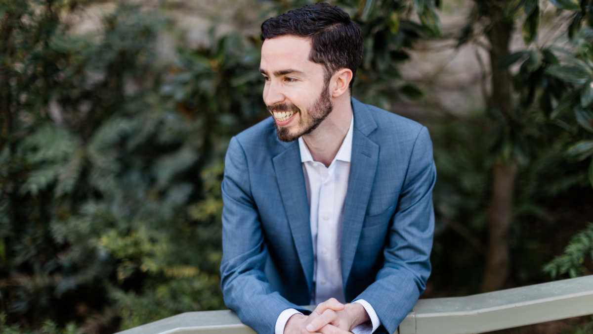 A man in a blue suit smiling brightly while leaning on a railing outdoors with green foliage in the background.