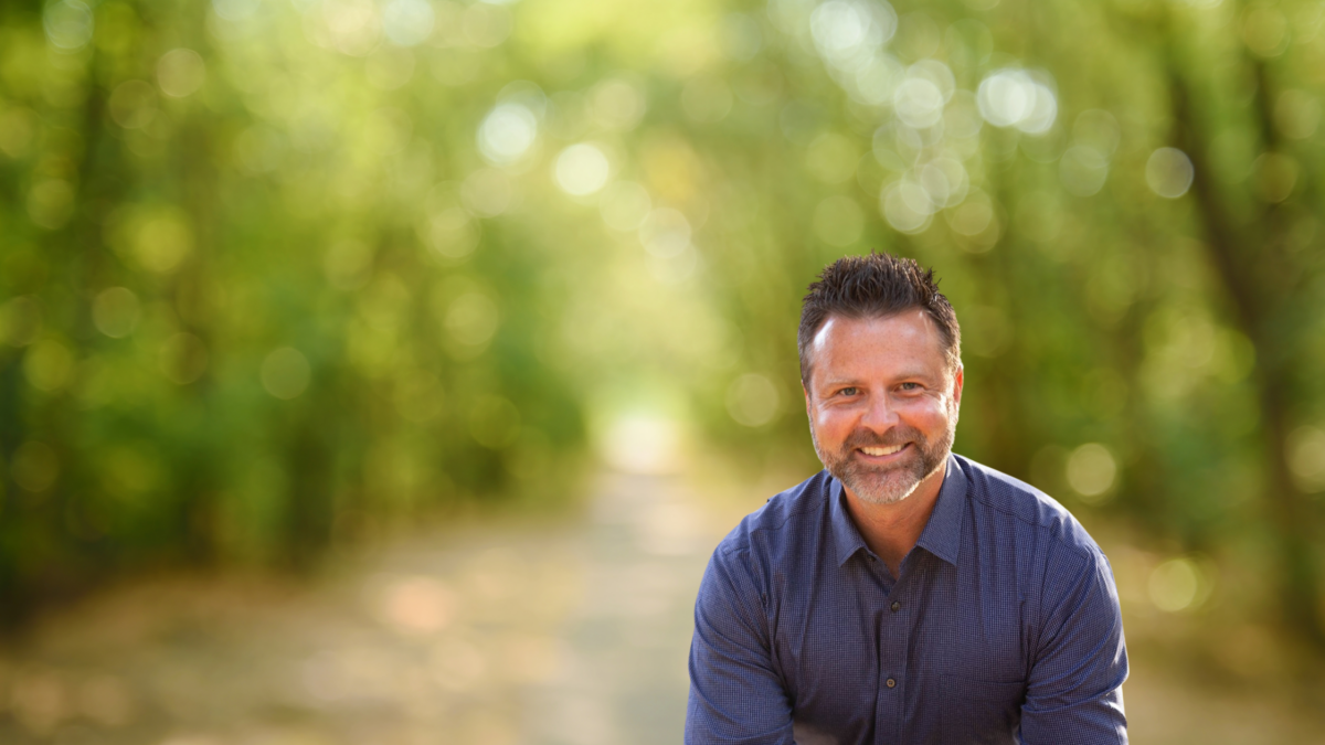 A smiling man with dark hair standing in a sunlit forested pathway with blurred greenery in the background.