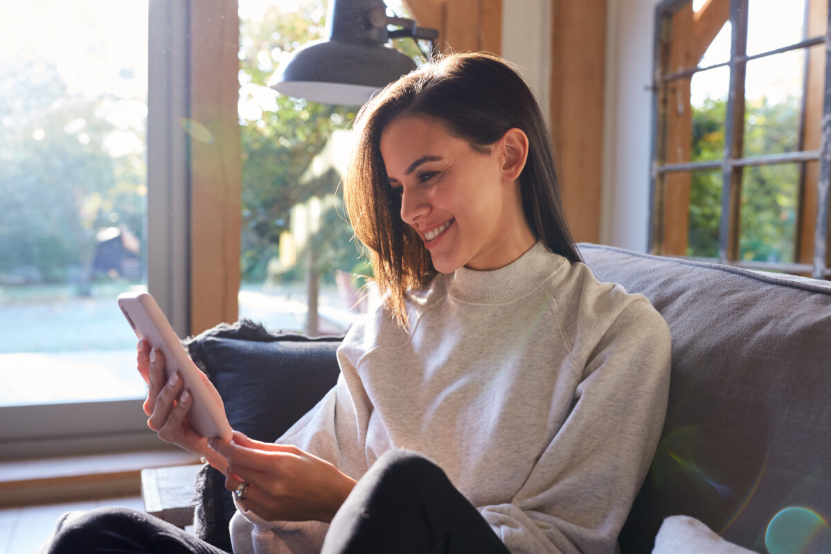 A woman relaxing on a couch by the window, enjoying the sunlight while smiling and using her tablet.