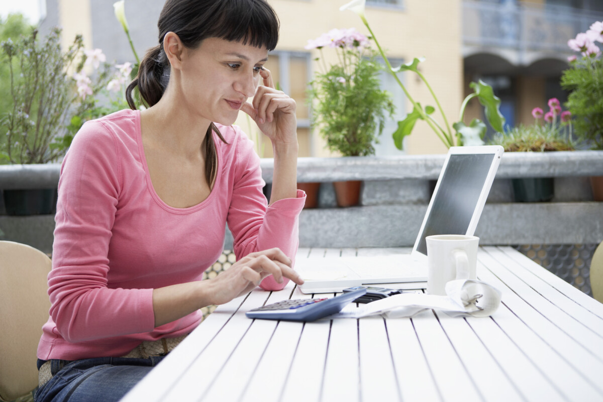 A woman in a pink top working intently on a calculator with a laptop open in front of her on an outdoor patio, possibly managing finances or budgeting. a cup of coffee sits beside her, suggesting a relaxed work setting.