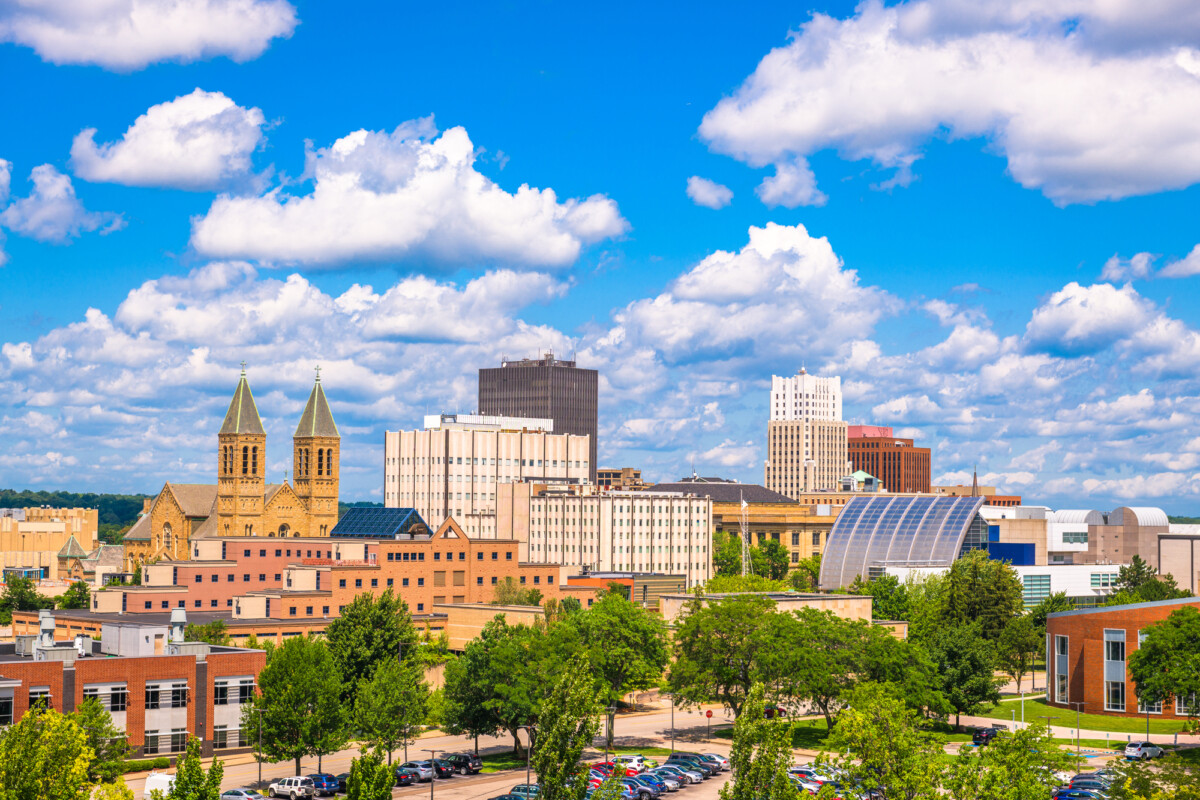 A sunny cityscape featuring a blend of architectural styles from modern buildings to historic church spires under a blue sky with fluffy clouds.