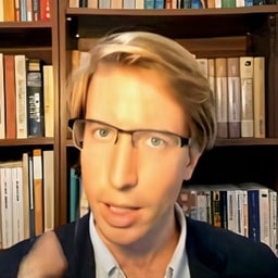 A person with blonde hair and glasses sitting in front of a bookshelf during a video call, possibly engaged in a virtual meeting or webinar.