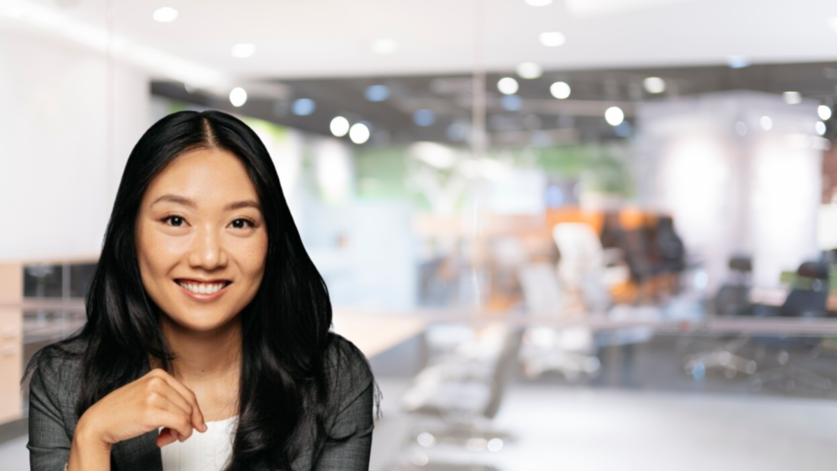 A woman with long black hair and a gray blazer smiles at the camera. She is seated with a hand resting on her chin. The background shows a brightly lit, modern office with glass walls and blurred details of desks and chairs.