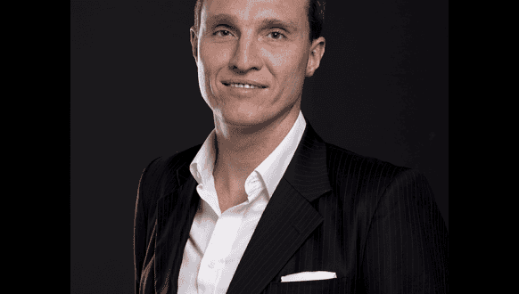A man in a dark pinstripe suit and white dress shirt stands against a dark background, smiling slightly at the camera.