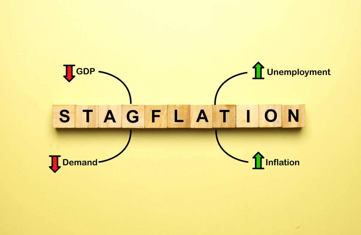 Wooden tiles spell “stagflation” on a yellow background. Arrows and labels show decreasing GDP and demand, and increasing unemployment and inflation, explaining the concept visually.