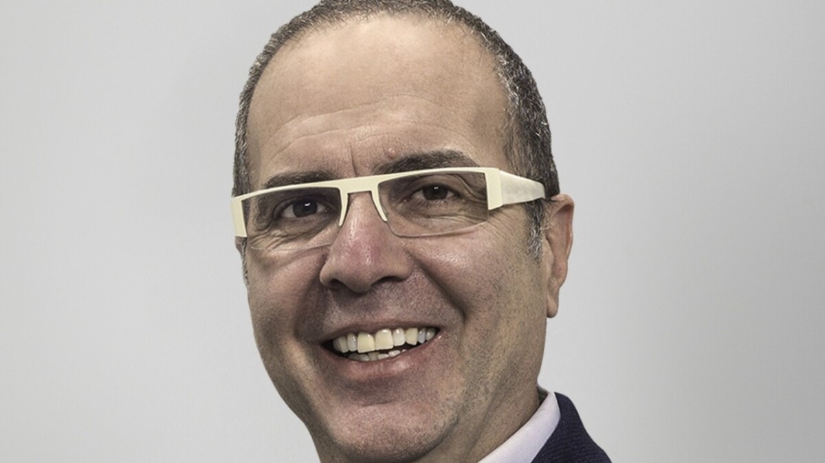A smiling man with short hair, wearing glasses with light-colored frames, and a dark suit jacket over a light shirt, against a plain light background.