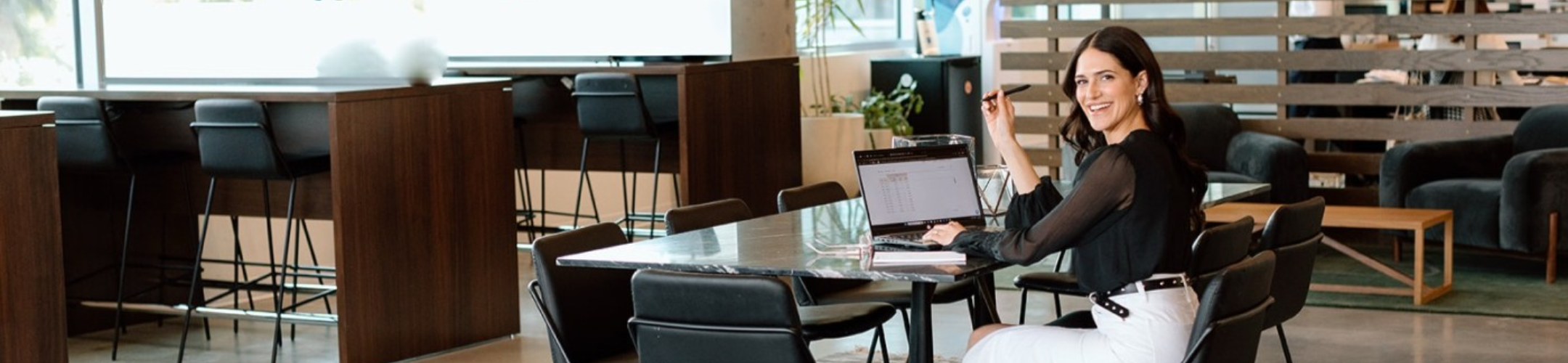 A woman in business attire sits at a marble table in a modern office space, smiling at the camera while working on a laptop with documents and a pen in hand.