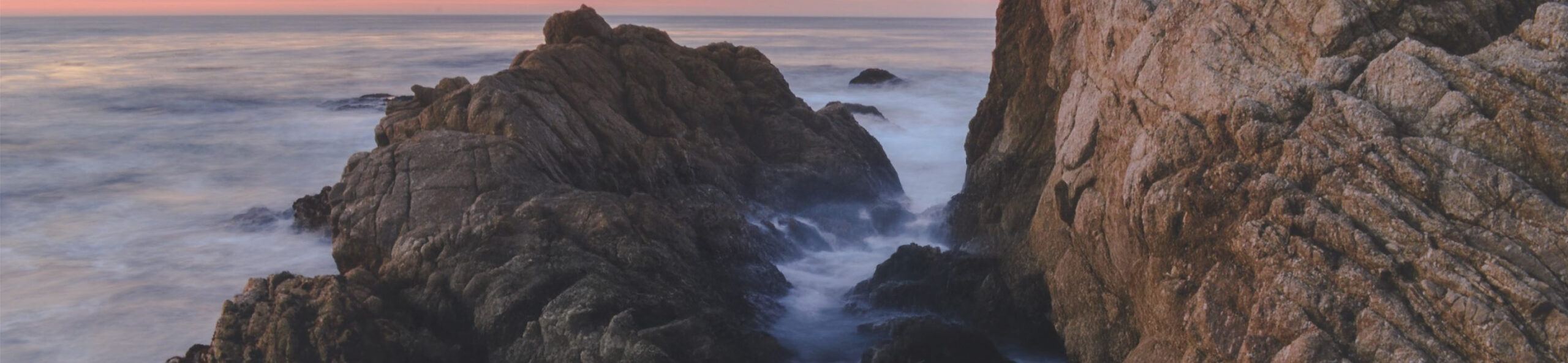 Rocky cliffs meet the ocean at sunset, with waves crashing against the rocks and a soft pink-orange glow in the sky.