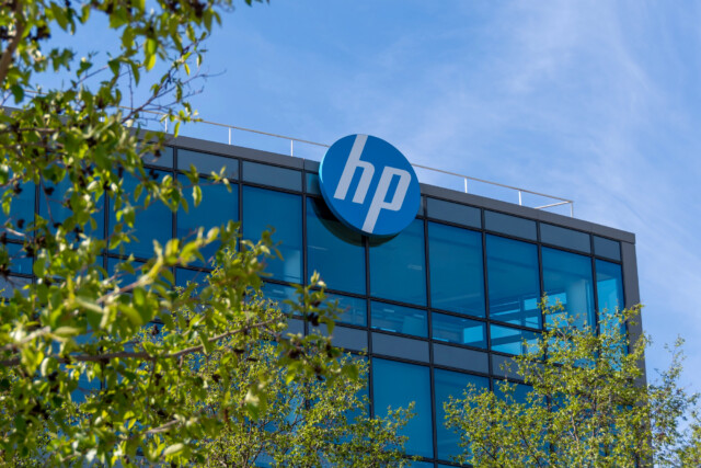 The exterior of an HP office building with large blue-tinted windows, a prominent circular hp logo at the top, and green tree branches in the foreground under a blue sky.