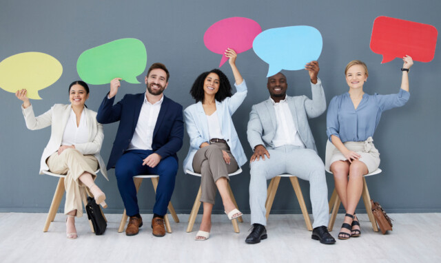 Five professionally dressed people sit in chairs, smiling and holding up colorful, blank speech bubble signs above their heads against a gray wall, illustrating the concept of clients of financial advisors sharing their online reviews visibly.
