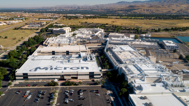 Aerial view of a large Micron Technology industrial facility with multiple white-roofed buildings, parking lots, and surrounding landscape; mountains are visible in the distance.