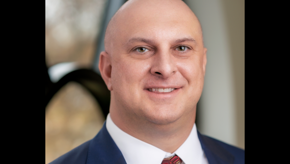 A bald man in a blue suit and red patterned tie smiles at the camera, with a blurred background showing a window and soft outdoor light.