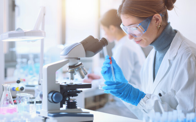 A scientist in a lab coat and safety goggles uses a pipette to transfer liquid into a petri dish next to a microscope.