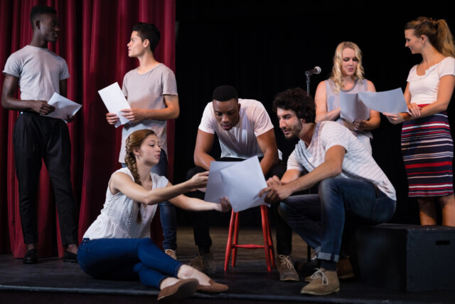 A group of actors on stage holding scripts and practicing lines together.